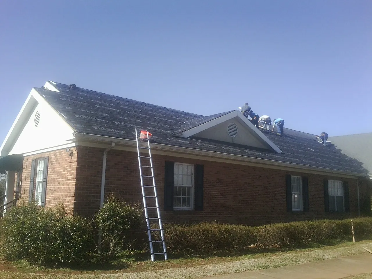 Crew tearing shingles off a brick ranch roof with a ladder set — Emergency Roof Repair work by our crew in Huxley
