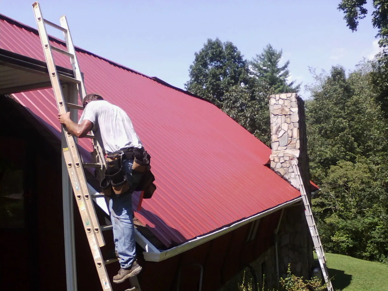 Roofer on a ladder beside a red metal roof and stone chimney — Soffit & Fascia Repair by Roofing Brothers in Huxley
